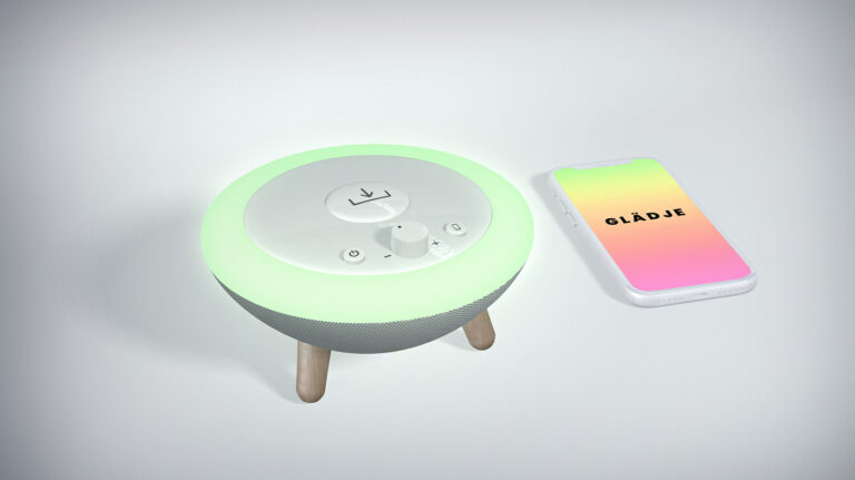 The concept Glädje. Glädje has the shape of a half sphere with the flat side facing upwards. It has a ring which lights up in different colours. It has three wooden legs. At the top there are some buttons for recieving a memory, turn on/off, volume, and sync with mobile. Next to Glädje is a smartphone with the Glädje app installed.